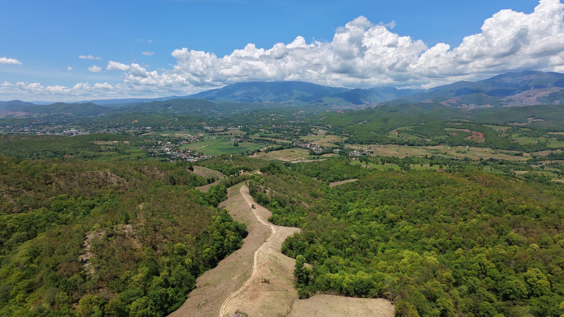 Aerial of a singletrack ridge trail dropping into the Mae Wang headwaters with Doi Inthanon on the horizon.